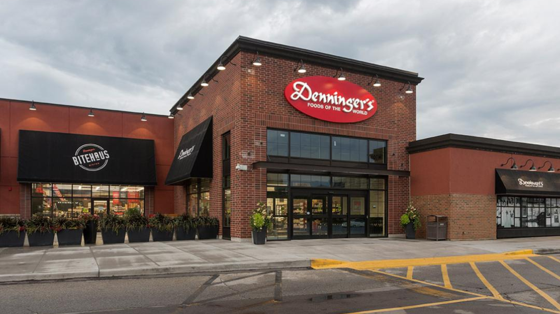 A Denninger’s grocery store exterior with a brick facade and a red “Denninger’s Foods of the World” sign above the main entrance. Large glass doors, storefront windows, and planters line the sidewalk under an overcast sky.
